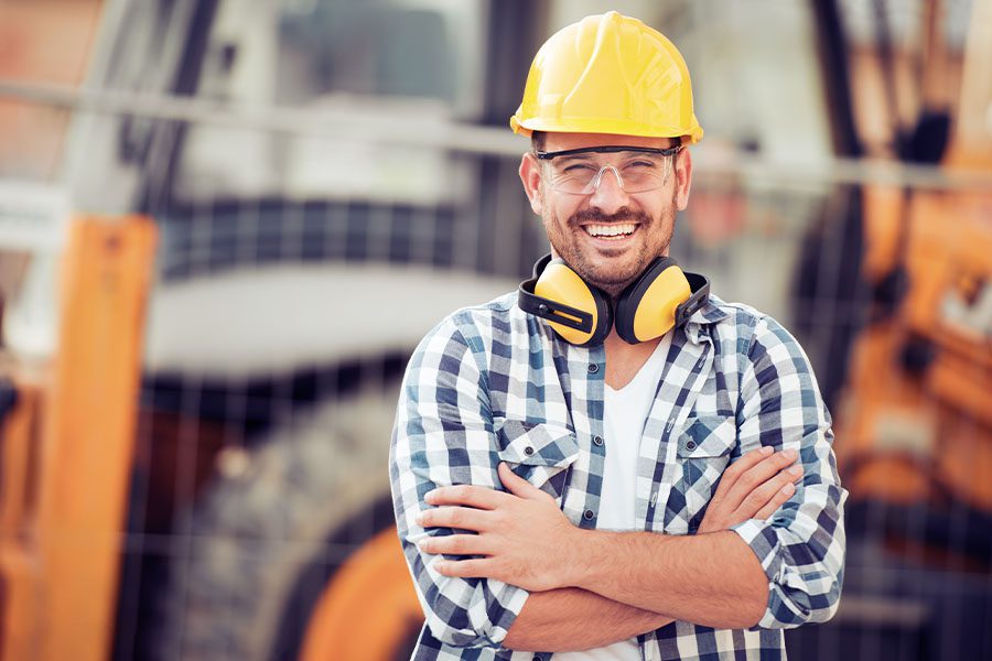 Specialized Business Insurance - Smiling Construction Worker in a Hard Hat Standing Against Large Equipment at a Construction Site Blurred in the Distance
